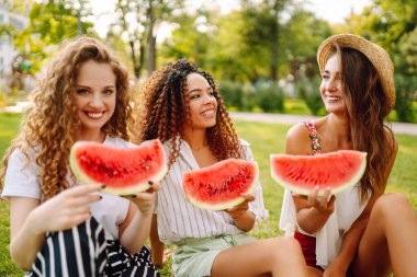 Üç genç kadın çimenlerde dinleniyor ve karpuz yiyor. İnsanlar, yaşam tarzı, seyahat, doğa ve tatil kavramı.