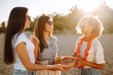 Üç genç kadın sahilde birlikte vakit geçiriyorlar piknik yapıyorlar, pizza yiyorlar. Fast food konsepti. Plaj tatili ve yaz tatili konsepti.