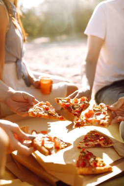 Elleri pizza dilimlerini yakından görüyor. Arkadaşlar plajda pizza yiyorlar. Fast food konsepti. Plaj tatili ve yaz tatili konsepti.