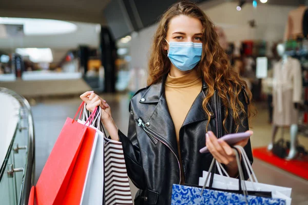 Telefonunu kullanarak alışveriş torbaları taşıyan koruyucu tıbbi maskeli genç bir kadın. Satın almalar, kara cuma, indirimler, satış konsepti. Çevrimiçi alışveriş kavramı.