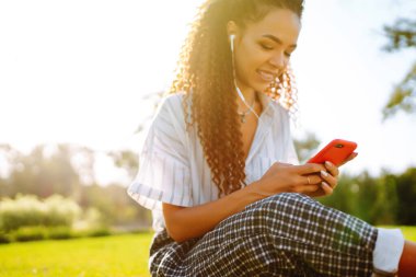 Parktaki yeşil çimlerde oturan genç bir kadın. Müzik dinleyen güzel bir kadın ya da kulaklıklı bir sesli kitap. Mobil iletişim konsepti. Güzel bir kadın dışarıda dinleniyor..