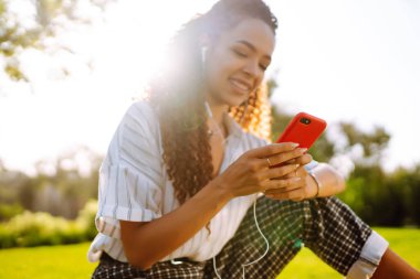 Parktaki yeşil çimlerde oturan genç bir kadın. Müzik dinleyen güzel bir kadın ya da kulaklıklı bir sesli kitap. Mobil iletişim konsepti. Güzel bir kadın dışarıda dinleniyor..