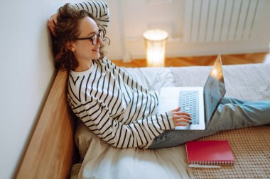 Eğitim ve online çalışma. Rahat ev içinde rahat bir yatakta oturan ve modern dizüstü bilgisayar üzerinde uzaktan çalışan genç bir bayan serbest yazar. Web mağazasında online alışveriş.