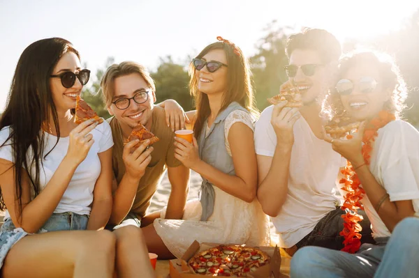 Group of young friends eating pizza, toasting with beerus and have fun on the beach. Happy friends resting together sitting near the sea. Fast food concept. Summer holidays, vacation, relax.
