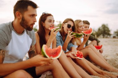 Genç arkadaşlar sahilde dinleniyor ve karpuz yiyorlar. Bir grup insan birlikte yaz partisinden zevk alır. İnsanlar, yaşam tarzı, seyahat, doğa ve tatil kavramı.
