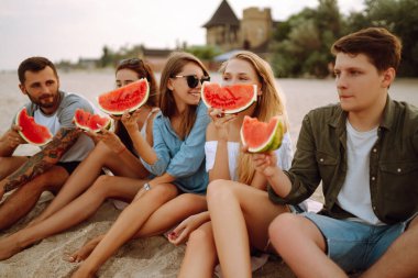 Genç arkadaşlar sahilde dinleniyor ve karpuz yiyorlar. Bir grup insan birlikte yaz partisinden zevk alır. İnsanlar, yaşam tarzı, seyahat, doğa ve tatil kavramı.