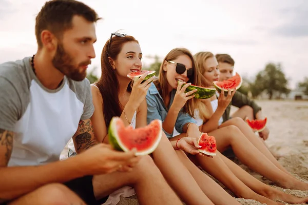 Genç arkadaşlar sahilde dinleniyor ve karpuz yiyorlar. Bir grup insan birlikte yaz partisinden zevk alır. İnsanlar, yaşam tarzı, seyahat, doğa ve tatil kavramı.