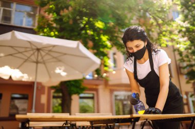 Koronavirüs salgını nedeniyle masaları dezenfekte eden genç bir garson. COVID-19.
