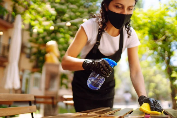 年轻的女服务员带着防护口罩和手套在餐桌上消毒 这是由于大肠病毒的流行 Covid — 图库照片