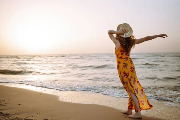 Young woman in beautiful dress and hat stands on Sea beach during an amazing sunset. Travel, weekend, relax and lifestyle concept.