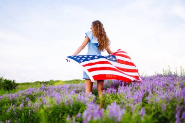 Çiçekli çayırda Amerikan bayrağı taşıyan genç bir kadın. 4 Temmuz. Bağımsızlık Günü. Vatansever bayram. ABD bayrağı rüzgarda dalgalanıyor.