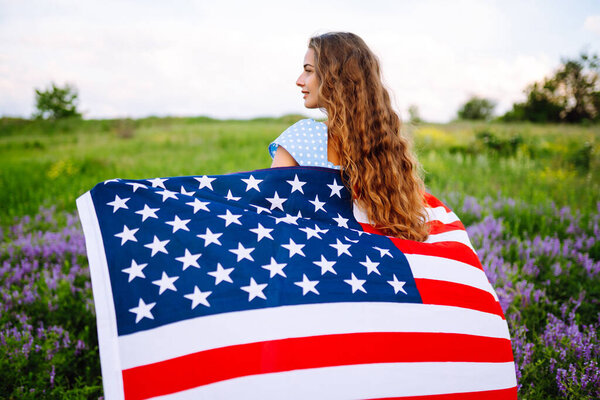 Young woman with american flag on blooming meadow. 4th of July. Independence Day. Patriotic holiday. USA flag fluttering in the wind.