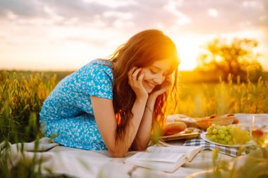 Güzel bir kadın battaniyeye oturmuş kitap okuyor ve dışarıda gülümsüyor. İnsanlar, yaşam tarzı, rahatlama ve tatil konsepti. Meyveli, peynirli, ekmekli piknik seti.