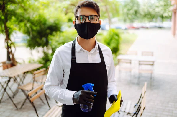 ラグとスプレーで保護マスクと手袋のウェイターは カフェの夏のテラスでテーブルを消毒します コロナウイルスのパンデミック時の食品配達人 Covid — ストック写真