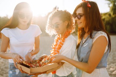 Üç genç kız arkadaş sahilde eğleniyor ve pizza yiyorlar. Fast food konsepti. Yaz tatili, tatiller, seyahat, yaşam tarzı ve insanlar konsepti