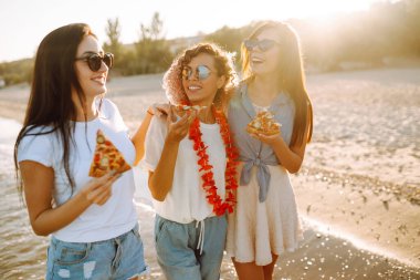 Üç genç kız arkadaş sahilde eğleniyor ve pizza yiyorlar. Fast food konsepti. Yaz tatili, tatiller, seyahat, yaşam tarzı ve insanlar konsepti