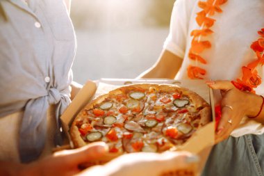 Elleri pizza dilimlerini yakından görüyor. Arkadaş grubu plajda pizza yiyor. Fast food konsepti. Sahilde piknik. Yaz tatili, tatiller, seyahat, yaşam tarzı ve insanlar konsepti.