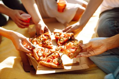Elleri pizza dilimlerini yakından görüyor. Arkadaş grubu plajda pizza yiyor. Fast food konsepti. Sahilde piknik. Yaz tatili, tatiller, seyahat, yaşam tarzı ve insanlar konsepti.