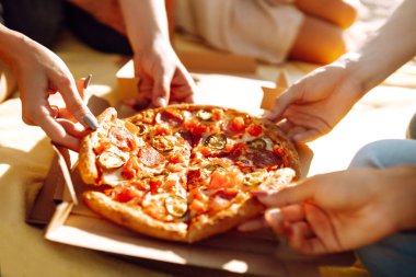 Elleri pizza dilimlerini yakından görüyor. Arkadaş grubu plajda pizza yiyor. Fast food konsepti. Sahilde piknik. Yaz tatili, tatiller, seyahat, yaşam tarzı ve insanlar konsepti.