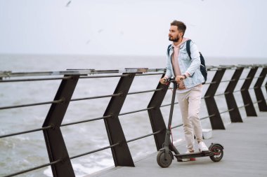Genç adam deniz kenarındaki bir rıhtımda elektrikli scooter kullanıyor. Ekolojik ulaşım kavramı.