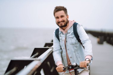 Genç adam deniz kenarındaki bir rıhtımda elektrikli scooter kullanıyor. Ekolojik ulaşım kavramı.