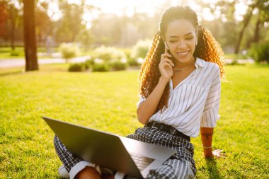 İletişim için dizüstü bilgisayar kullanan genç bir kadın. Parktaki çimlerin üzerinde oturan aile veya arkadaşlarıyla görüntülü konuşma. Video aramasında çevrimiçi toplantı. 
