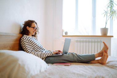 Genç bayan serbest çalışıyor ve evde karantinada çalışırken notlar alıyor. Telsiz kulaklıklı Freelancer laptoptan arıyor, web kamerasıyla konuşuyor. Eğitim, alışveriş ve online çalışma