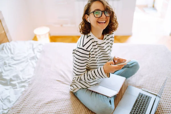 Young female freelancer works and makes notes while working at home in quarantine lockdown. Freelancer with wireless headphones calling on laptop, talk by webcam. Education,shopping and working online