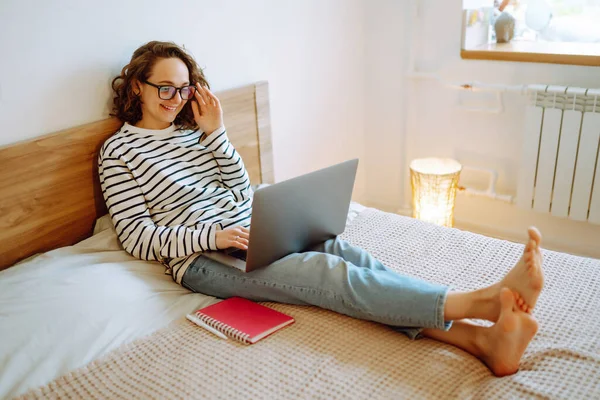 Young female freelancer works and makes notes while working at home in quarantine lockdown. Freelancer with wireless headphones calling on laptop, talk by webcam. Education,shopping and working online