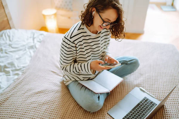 Young female freelancer works and makes notes while working at home in quarantine lockdown. Freelancer with wireless headphones calling on laptop, talk by webcam. Education,shopping and working online