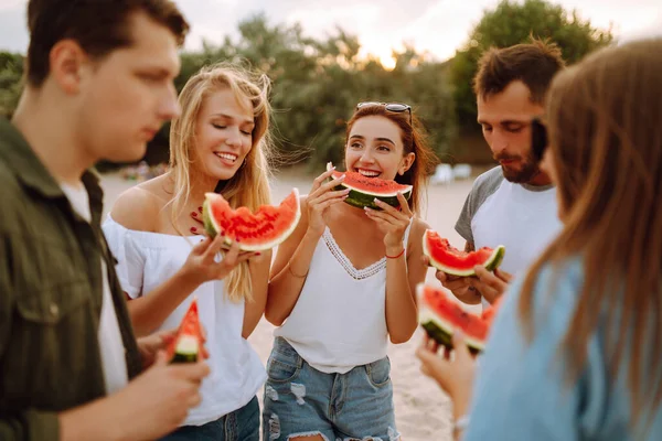 ピクニックをしたり スイカを食べたり ビーチで夏のパーティーを楽しむ若い友人のグループ 幸せな友人が一緒に海の近くに座って休んでいる ビーチホリデーと夏休みのコンセプト — ストック写真