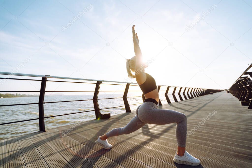 Young fitness woman exercising and stretching outdoors in the morning. Fit healthy athlete is doing workout at the beach pier. Sport, Active life.