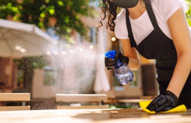 COVID-19 'u önlemek için dezenfekte ediyorum. Koruyucu yüz maskesi takmış genç bir garson ve dezenfektan spreyi ve kafede mikrofiber bezle eldiven temizleme masası..