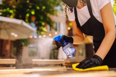 COVID-19 'u önlemek için dezenfekte ediyorum. Koruyucu yüz maskesi takmış genç bir garson ve dezenfektan spreyi ve kafede mikrofiber bezle eldiven temizleme masası..