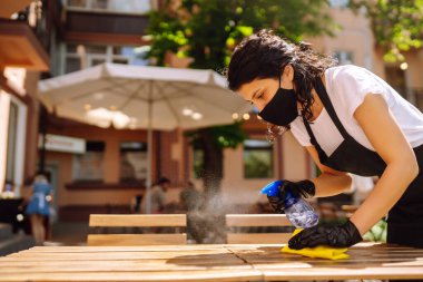 COVID-19 'u önlemek için dezenfekte ediyorum. Koruyucu yüz maskesi takmış genç bir garson ve dezenfektan spreyi ve kafede mikrofiber bezle eldiven temizleme masası..