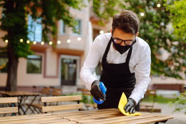 COVID-19 'u önlemek için dezenfekte ediyorum. Karantina şehrindeki kafede dezenfektan spreyi ve mikrofiber bezle koruyucu maske ve eldiven temizleme masasında garson..