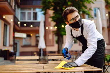 COVID-19 'u önlemek için dezenfekte ediyorum. Karantina şehrindeki kafede dezenfektan spreyi ve mikrofiber bezle koruyucu maske ve eldiven temizleme masasında garson..