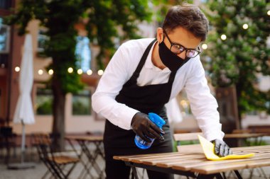 COVID-19 'u önlemek için dezenfekte ediyorum. Karantina şehrindeki kafede dezenfektan spreyi ve mikrofiber bezle koruyucu maske ve eldiven temizleme masasında garson..