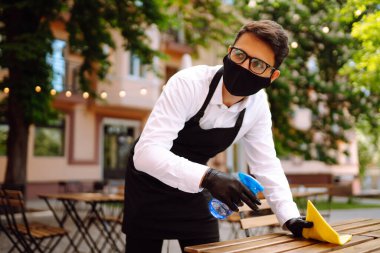 COVID-19 'u önlemek için dezenfekte ediyorum. Karantina şehrindeki kafede dezenfektan spreyi ve mikrofiber bezle koruyucu maske ve eldiven temizleme masasında garson..