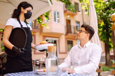 Koruyucu tıbbi maskeli ve eldivenli genç garson dışarıda yemek servisi yapıyor. Coronavirus salgını yaşam tarzı. Sosyal uzaklık.