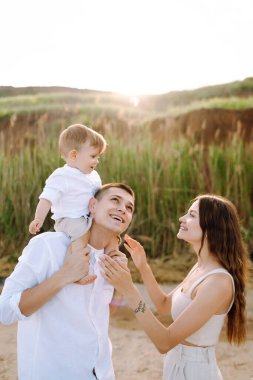 Küçük oğluyla sahilde vakit geçiren neşeli, genç bir aile. Baba, anne ve çocuk mavi denizin ve gökyüzünün arka planına karşı. Aile, çocukluk, aktif yaşam tarzı konsepti.