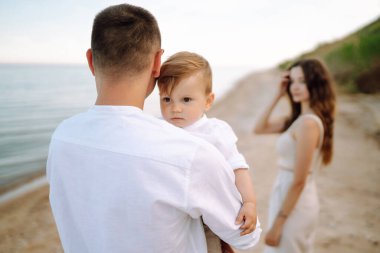Küçük oğluyla sahilde vakit geçiren neşeli, genç bir aile. Baba, anne ve çocuk mavi denizin ve gökyüzünün arka planına karşı. Aile, çocukluk, aktif yaşam tarzı konsepti.