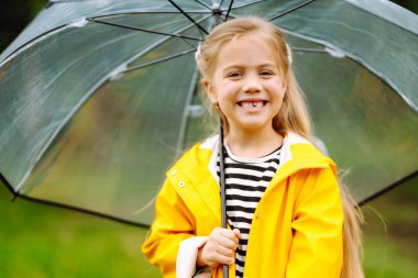  Şeffaf şemsiyeli komik kız yağmurdan hoşlanıyor. Sarı yağmurluk ve lastik çizme giyiyor. Küçük kız doğada oynuyor. Mutlu çocukluk kavramı. 
