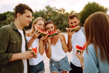 Genç arkadaşlar sahilde dinleniyor ve karpuz yiyorlar. Bir grup genç birlikte deniz kenarında oturuyor ve yaz partisinin tadını çıkarıyor. İnsanlar, yaşam tarzı, seyahat, doğa ve tatil kavramı.