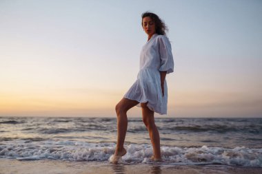 Beyaz kanat çırpan elbiseli genç ve mutlu bir kadın deniz kıyısında yürüyor. Kız sihirli gün doğumuna bakıyor. Yaz zamanı. Seyahat, hafta sonu, rahatlama ve yaşam tarzı konsepti.