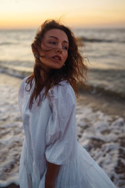 Beyaz kanat çırpan elbiseli genç ve mutlu bir kadın deniz kıyısında yürüyor. Kız sihirli gün doğumuna bakıyor. Yaz zamanı. Seyahat, hafta sonu, rahatlama ve yaşam tarzı konsepti.