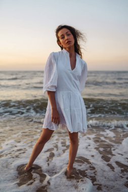 Beyaz kanat çırpan elbiseli genç ve mutlu bir kadın deniz kıyısında yürüyor. Kız sihirli gün doğumuna bakıyor. Yaz zamanı. Seyahat, hafta sonu, rahatlama ve yaşam tarzı konsepti.