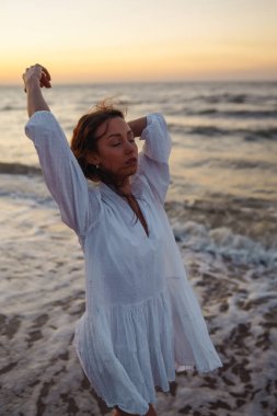 Beyaz kanat çırpan elbiseli genç ve mutlu bir kadın deniz kıyısında yürüyor. Kız sihirli gün doğumuna bakıyor. Yaz zamanı. Seyahat, hafta sonu, rahatlama ve yaşam tarzı konsepti.