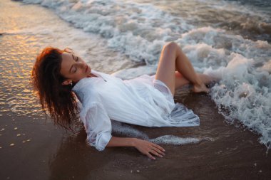 Beyaz kanat çırpan elbiseli genç ve mutlu bir kadın deniz kıyısında yürüyor. Kız sihirli gün doğumuna bakıyor. Yaz zamanı. Seyahat, hafta sonu, rahatlama ve yaşam tarzı konsepti.