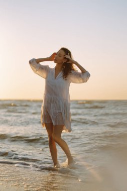 Beyaz kanat çırpan elbiseli genç ve mutlu bir kadın deniz kıyısında yürüyor. Kız sihirli gün doğumuna bakıyor. Yaz zamanı. Seyahat, hafta sonu, rahatlama ve yaşam tarzı konsepti.